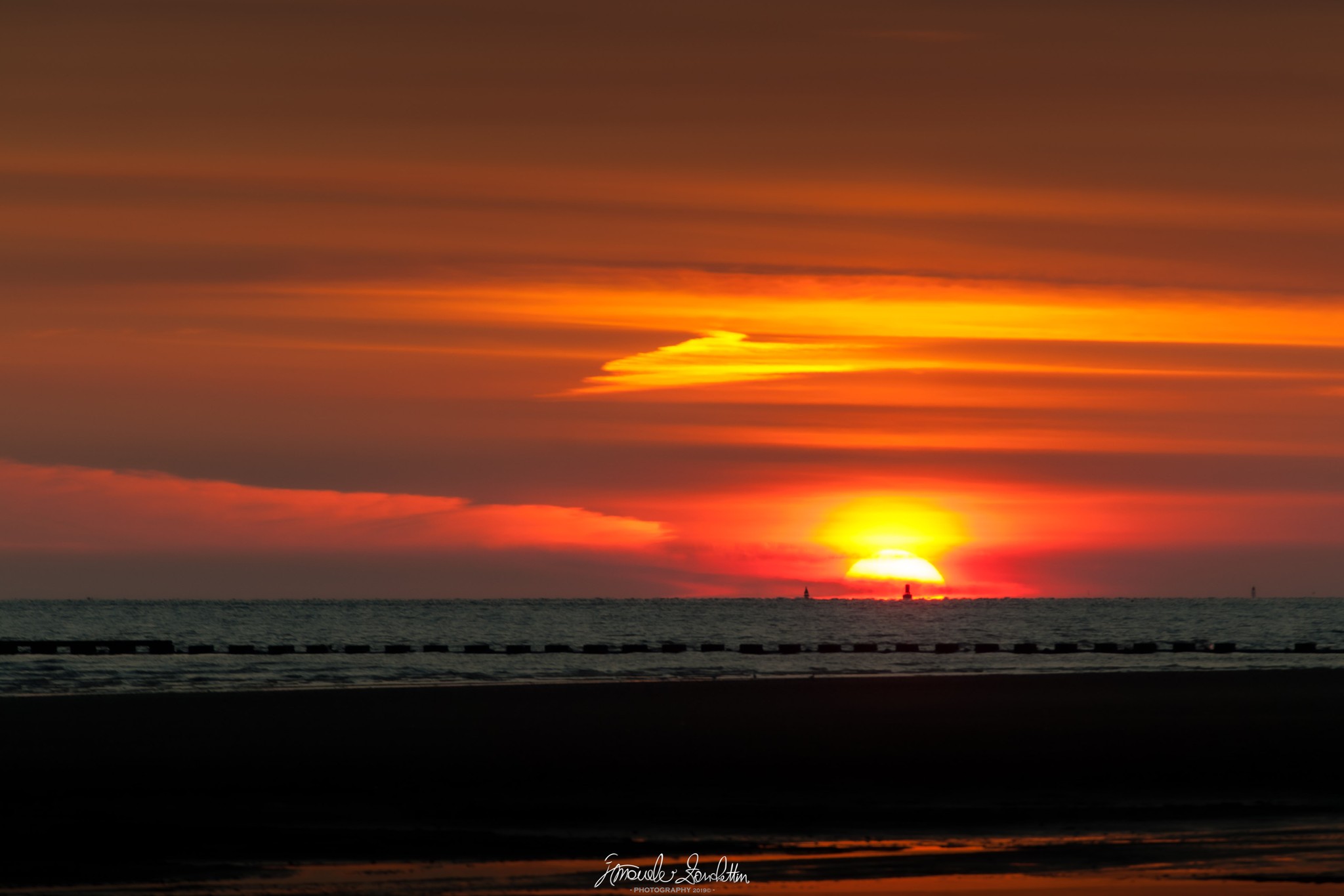 A low sun beneath orange clouds above the sea and a dark shoreline
